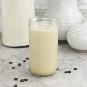 Tall glass with creamy protein shake inside, coffee beans scattered on marble bench top, white ceramic vases in background.