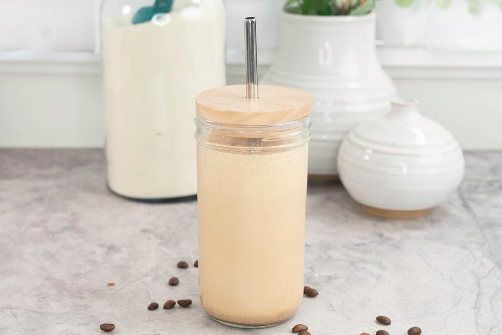 Tall glass with lid and metal straw with creamy protein shake inside, coffee beans scattered on marble bench top, white ceramic vases in background and large jar of protein.