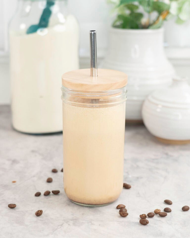 Tall glass with lid and metal straw with creamy protein shake inside, coffee beans scattered on marble bench top, white ceramic vases in background and large jar of protein.