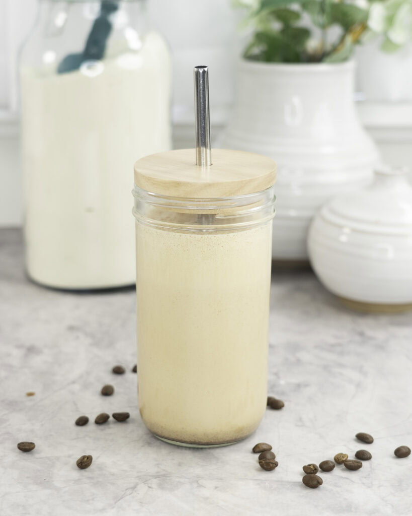Tall glass with lid and metal straw with creamy protein shake inside, coffee beans scattered on marble bench top, white ceramic vases in background and large jar of protein.