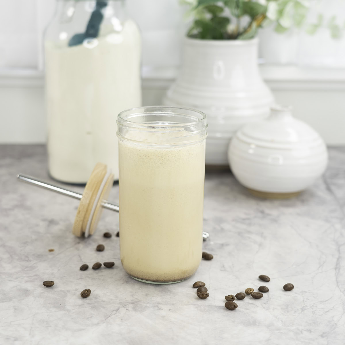 Tall glass with lid and metal straw sitting on benchtop with creamy protein shake inside, coffee beans scattered on marble bench top, white ceramic vases in background and large jar of protein.