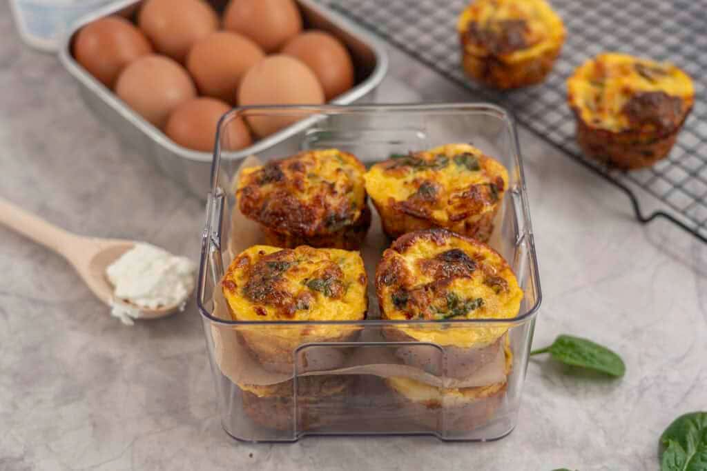 Eight egg muffins stacked in a quart container four on top of one another separated by baking paper, stingless steel container of eggs and metal baking tray in background with two egg muffins.