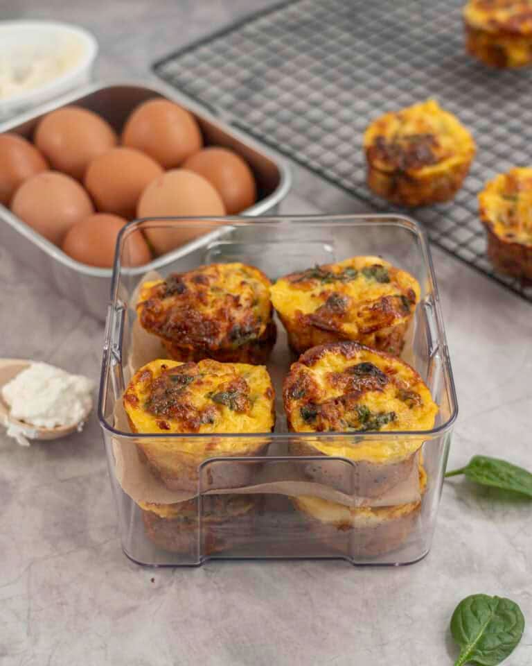 Eight egg muffins stacked in a quart container four on top of one another separated by baking paper, stingless steel container of eggs and metal baking tray in background with two egg muffins.