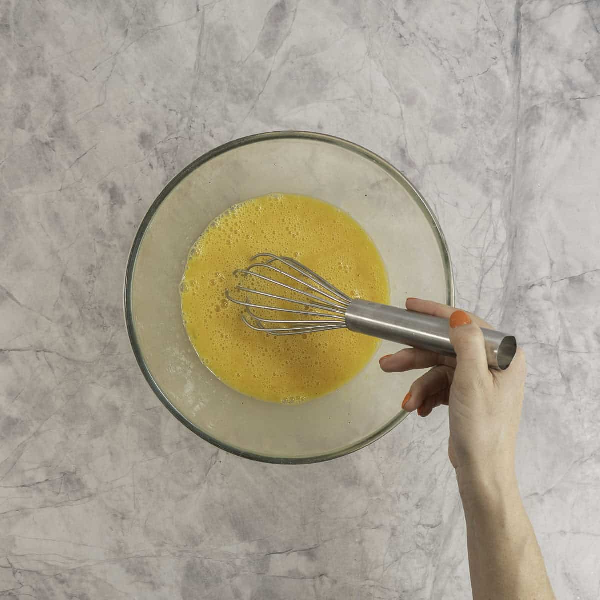 Womans hand holding a metal whisk stirring eggs in a large glass bowl.