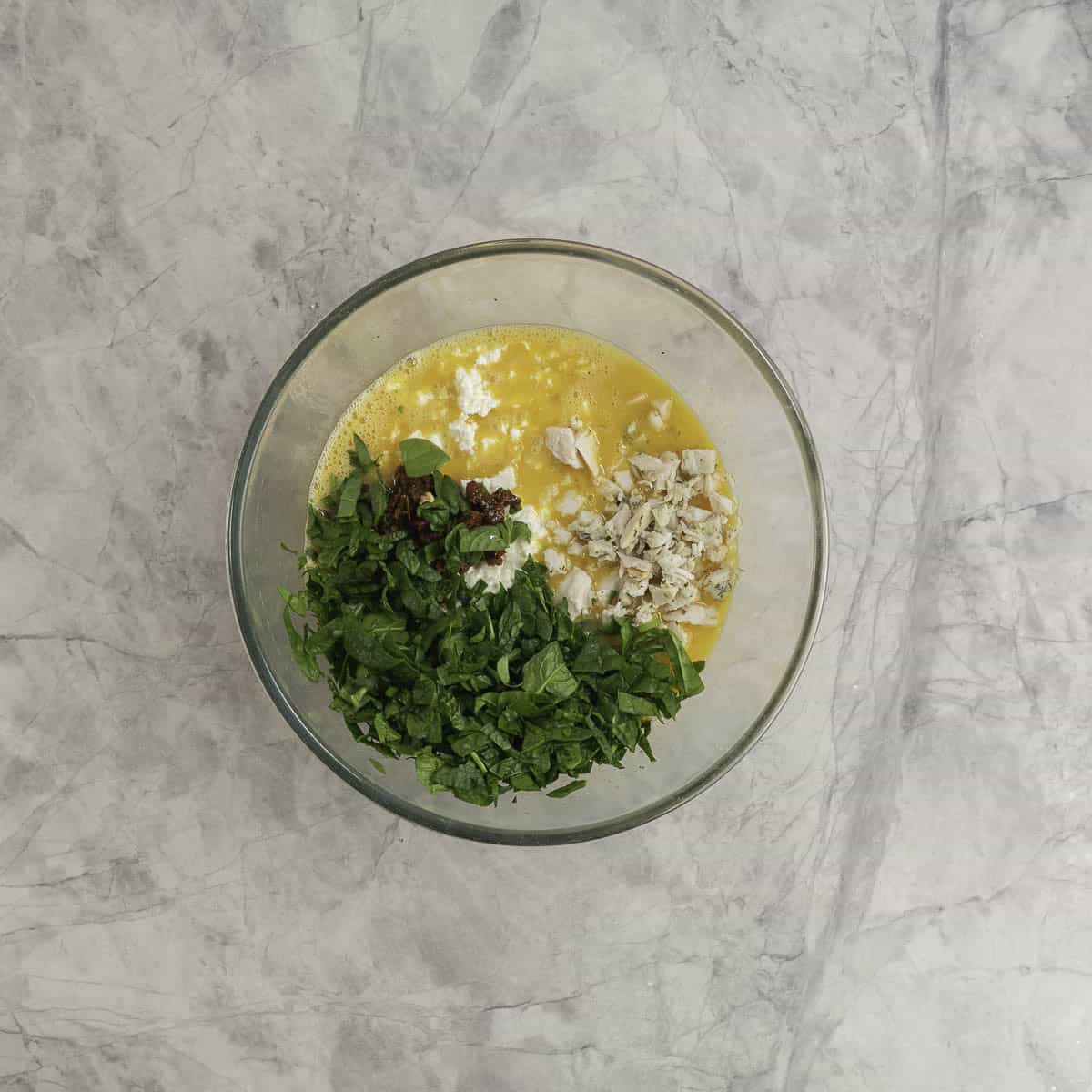 Chopped spinach, chicken, cottage cheese, sun-dried tomatoes added into whisked egg in large glass bowl on marble bench top.