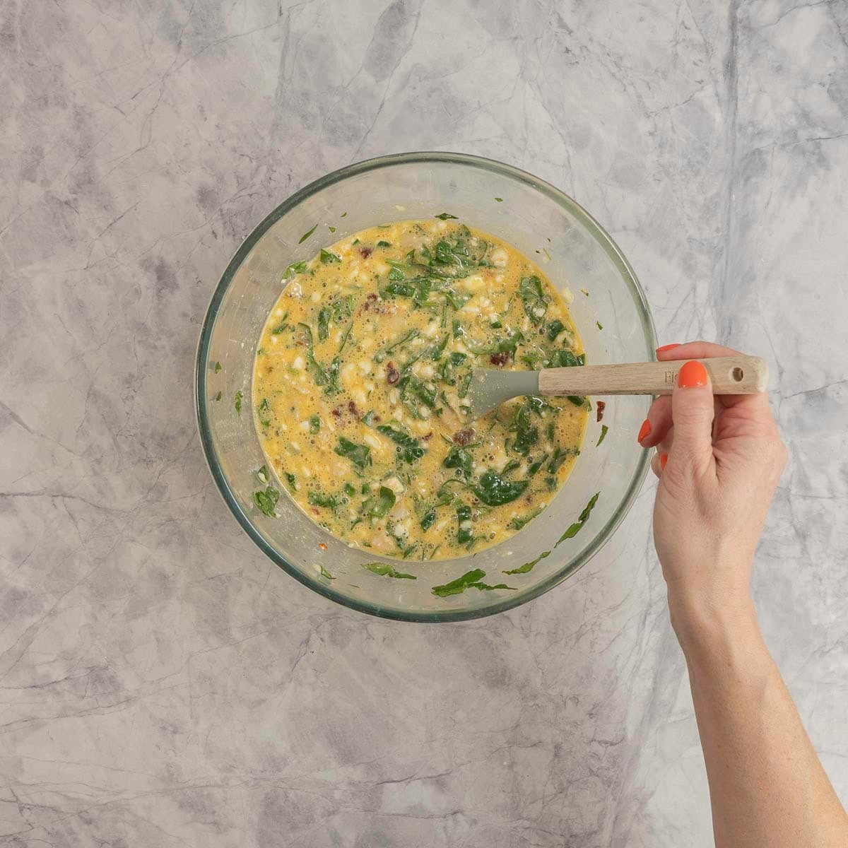 Ingredients in a large glass bowl on marble benchtop, hand holding a spatula stirring egg cottage cheese spinach mixture.