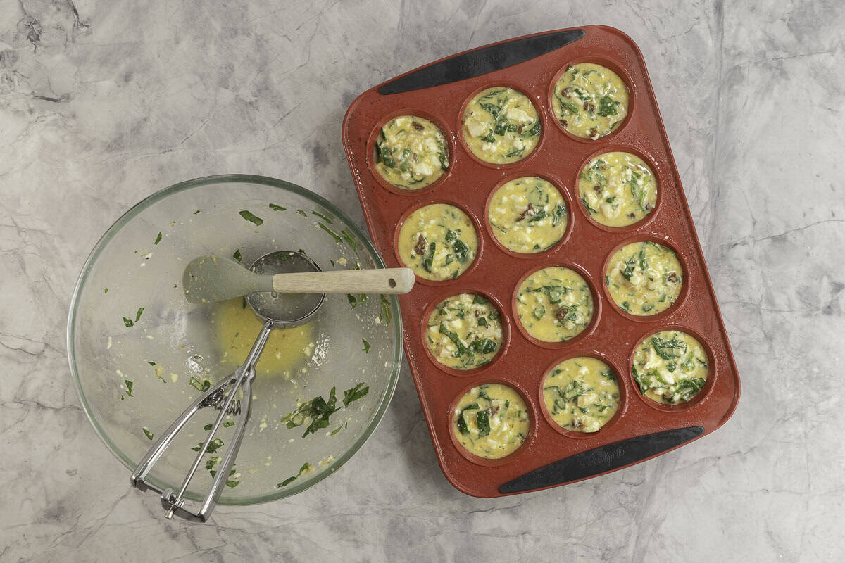 A red silicone muffin tray with egg mixture portioned inside, large glass bowl empty with scoop and spatula inside.