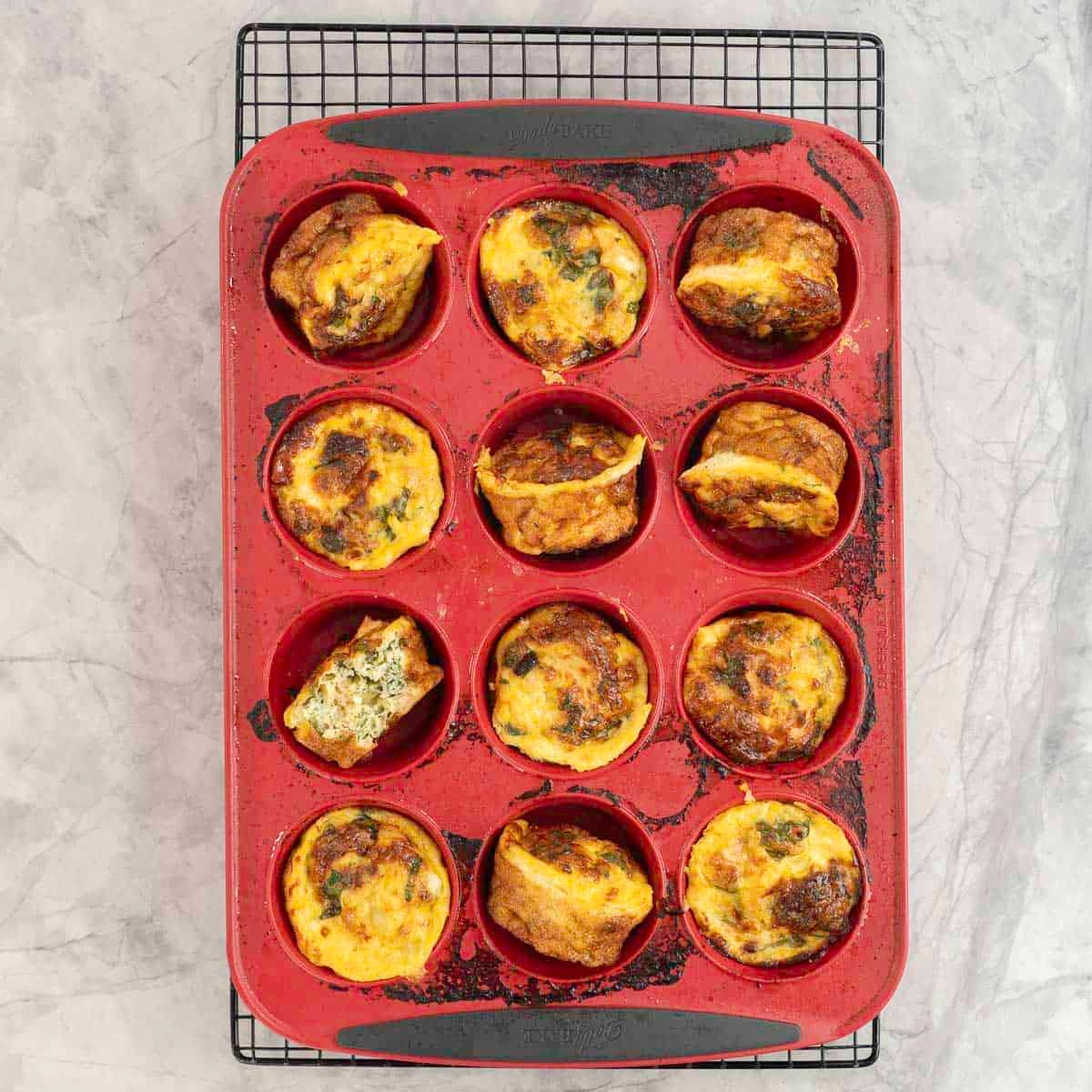 Cooked egg muffins inside a red silicone muffin tray placed on a cooling rack, muffins turned onto their side for removal.