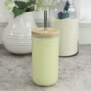 Tall glass with lid and metal straw with green protein shake inside on marble bench top, white ceramic vase in background with greenery and large jar of protein.