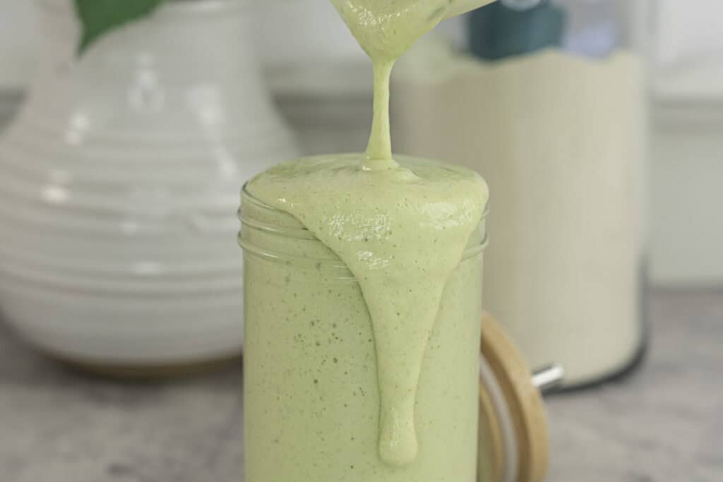 Tall glass with green protein shake being poured to overflow on marble bench top, white ceramic vase in background with greenery and large jar of protein.
