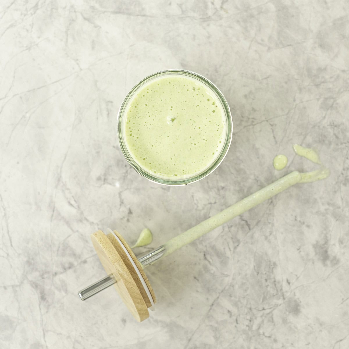 Tall glass with green smoothie inside, metal straw taken out with green smoothie dripping on marble bench top.