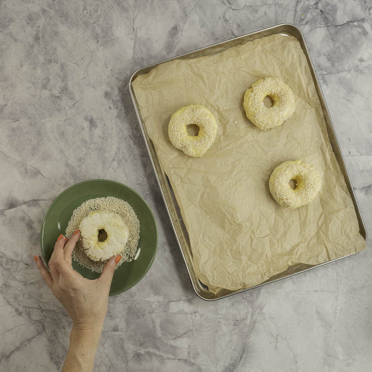 Metal tray lined with baking paper with three bagels dipped on top side with sesame seeds, green plate with sesame seeds on it, woman's hand holding a bagel and dipping topside in.