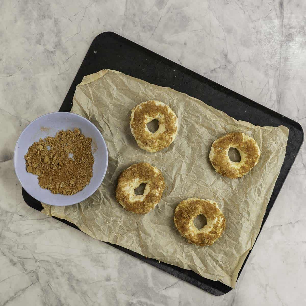 Metal baking tray lined with baking paper, four bagels topped with brown sugar mixture, blue bowl with brown sugar mixture inside.