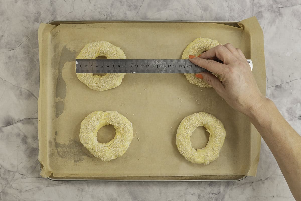 Four bagels on metal tray lined with baking paper, woman's hand holding a metal ruler measuring the size if whole in the middle.