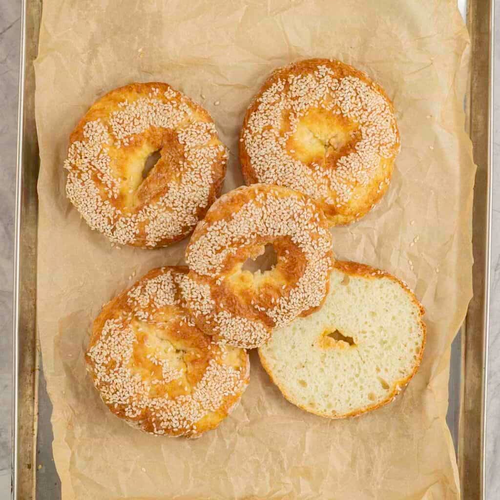 Cooked bagels with small holes in the centre topped with sesame seeds on metal tray lined with baking paper.