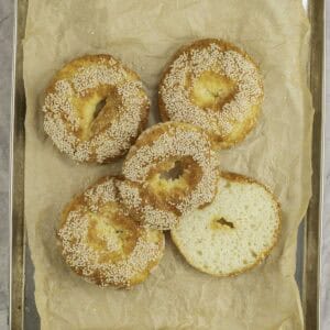 Cooked bagels with small holes in the centre topped with sesame seeds on metal tray lined with baking paper.