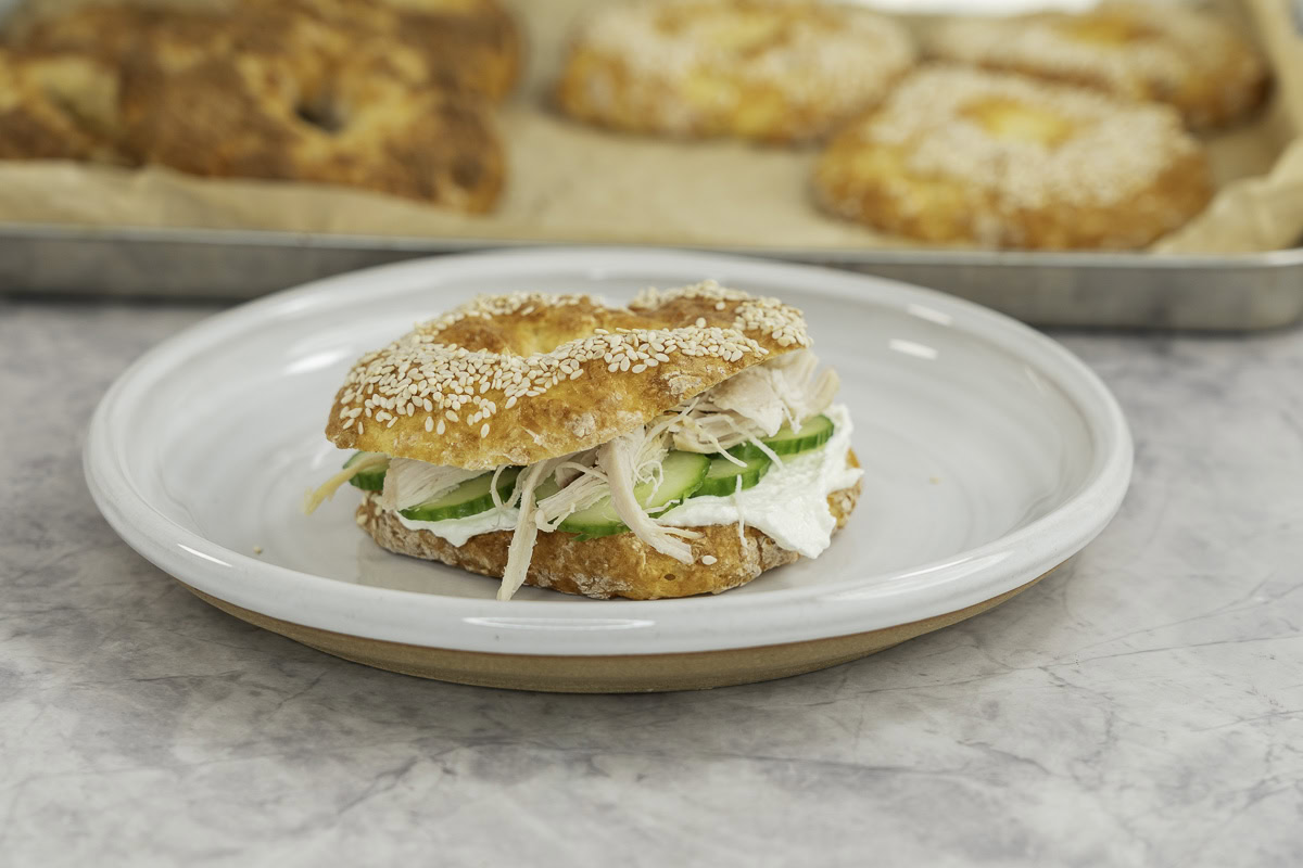 A bagel on a plate cut open with cream cheese cucumber slices and fresh chicken inside.