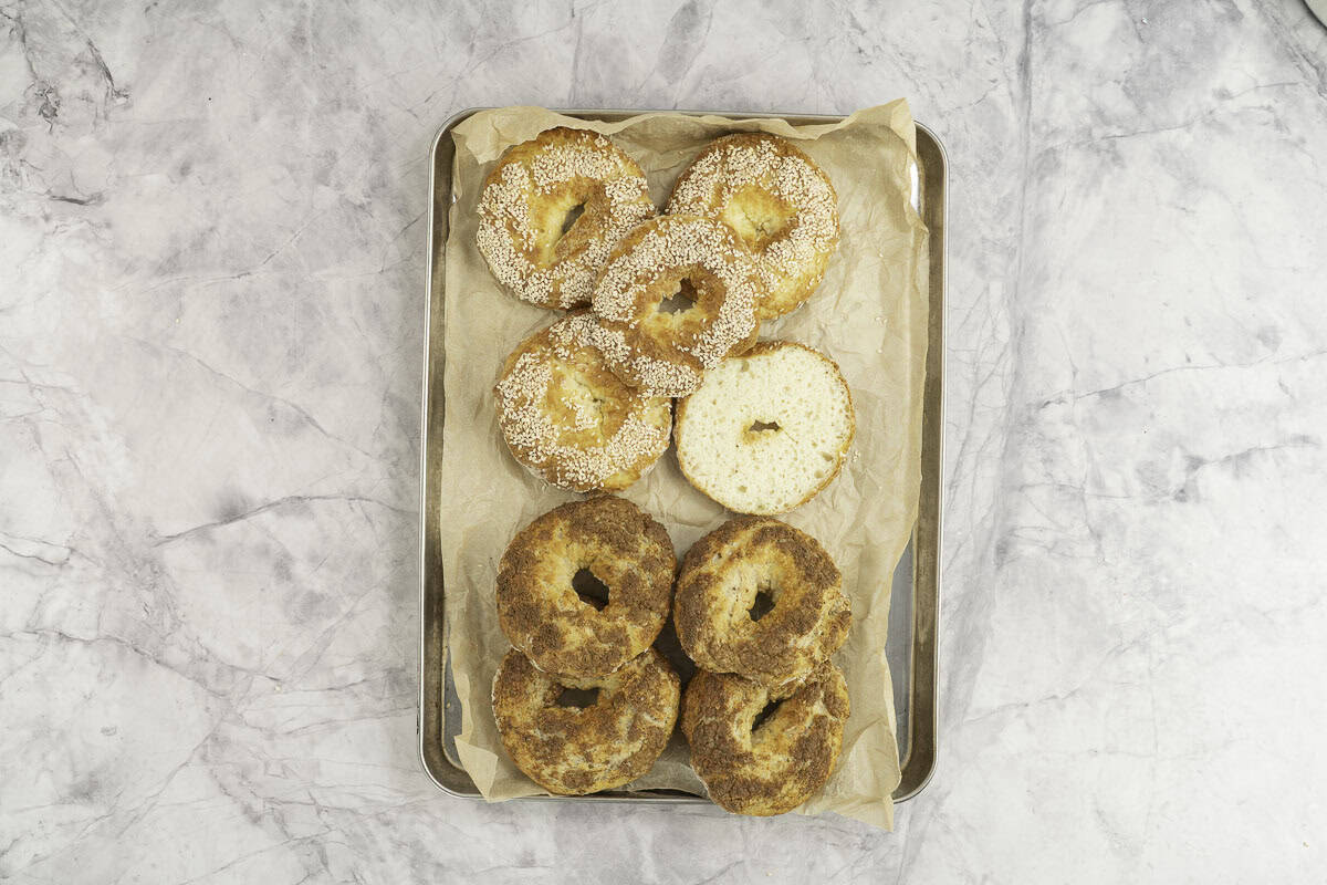 Eight cooked bagels on metal baking tray lined with baking paper, four topped with seasme seeds, four with brown sugar mixture.