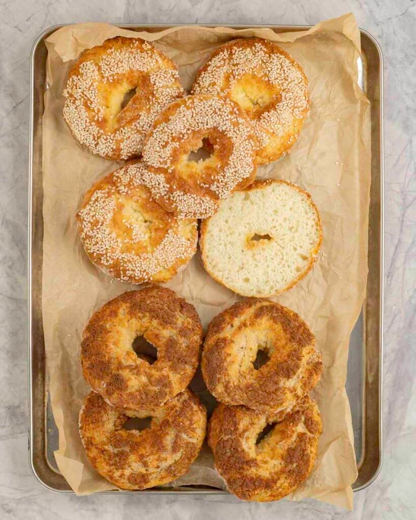 Eight cooked bagels on metal baking tray lined with baking paper, four topped with seasme seeds, four with brown sugar mixture.
