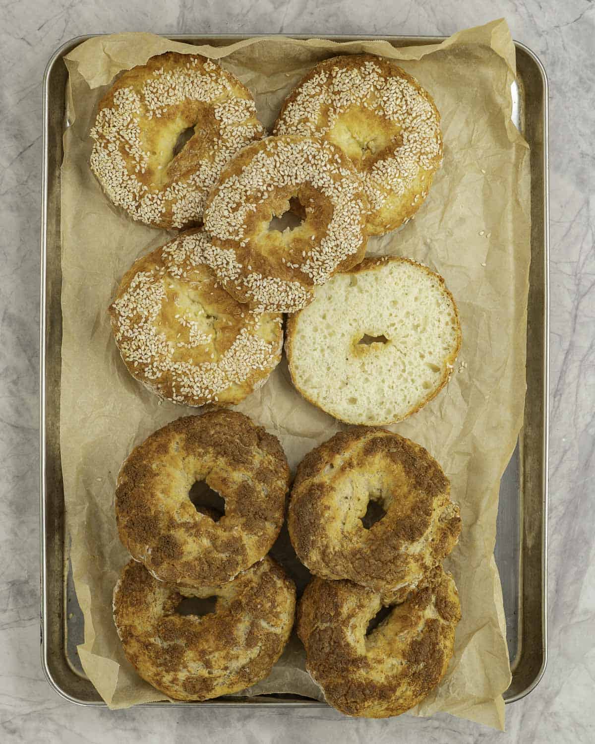 Eight cooked bagels on metal baking tray lined with baking paper, four topped with seasme seeds, four with brown sugar mixture.