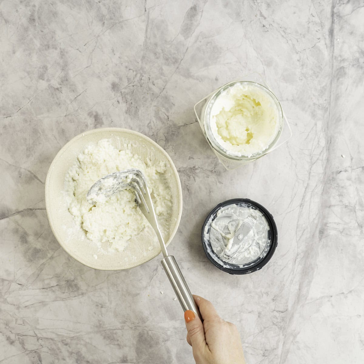 Hand holding a potato masher in a bowl of cottage cheese, smoothie cup with blender lid on benchtop with cottage also.