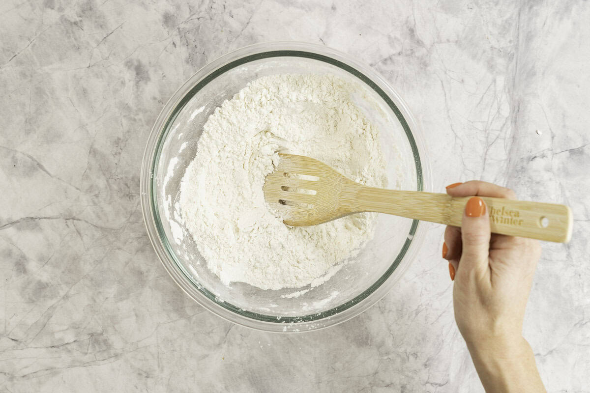 Hand holding a wooden spoon mixing flour and dry ingredients in glass bowl.