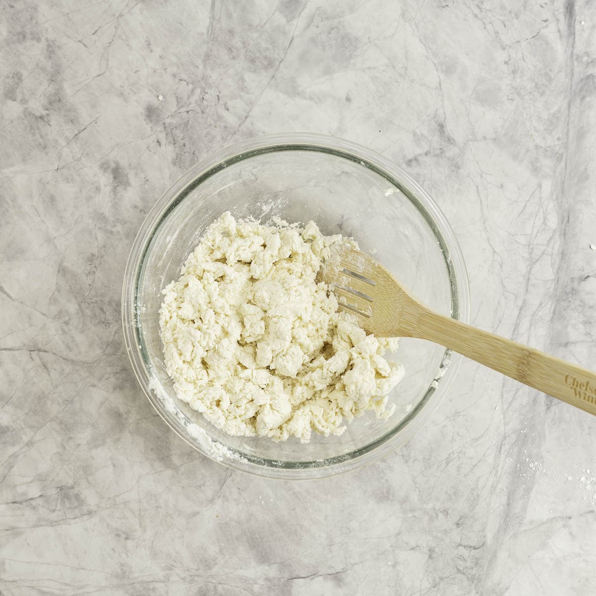 Wooden spoon mixing bagel mixture together in large glass bowl on marble benchtop.