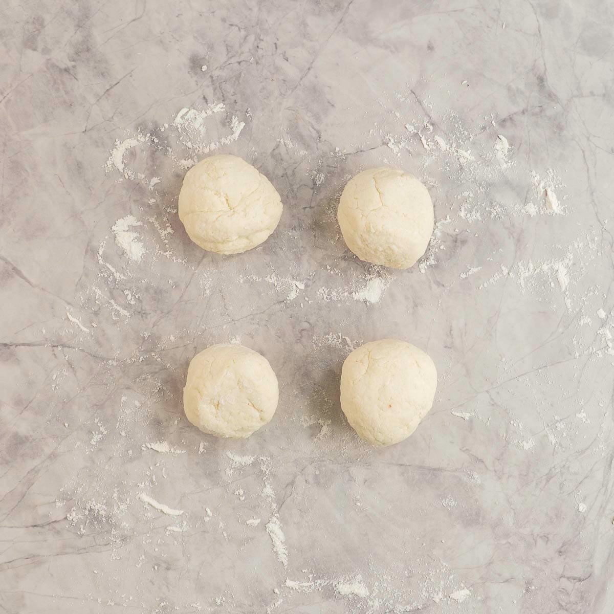 Four balls of bagel dough on marble benchtop scattered lightly with flour.