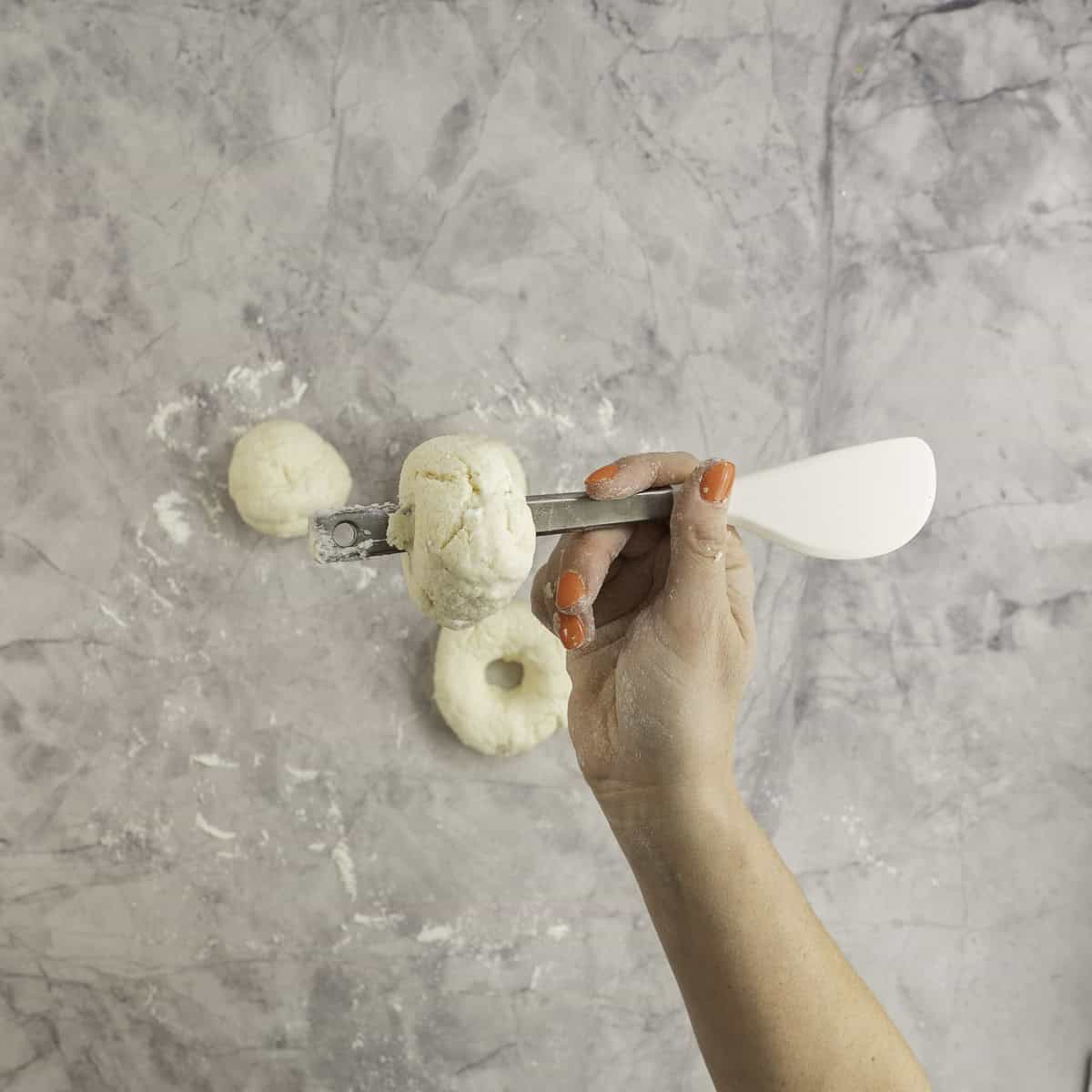 Hand holding a metal spatula handle horizontally with bagel dough pierced through forming a hole in the middle.
