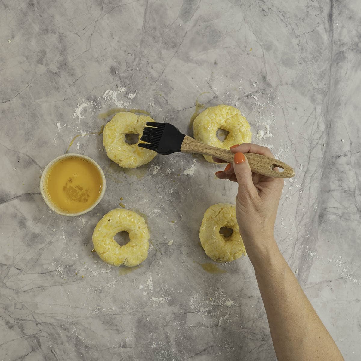 Four bagel shaped dough on marble benchtop scattered lightly with flour, hand holding a basting brush basting bagels with whisked egg.
