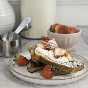 A stack of french toast with yogurt on top, cut strawberries, icing sugar and maple syrup. Bowl of fresh strawberries in small blush pink dish in background and white vases.