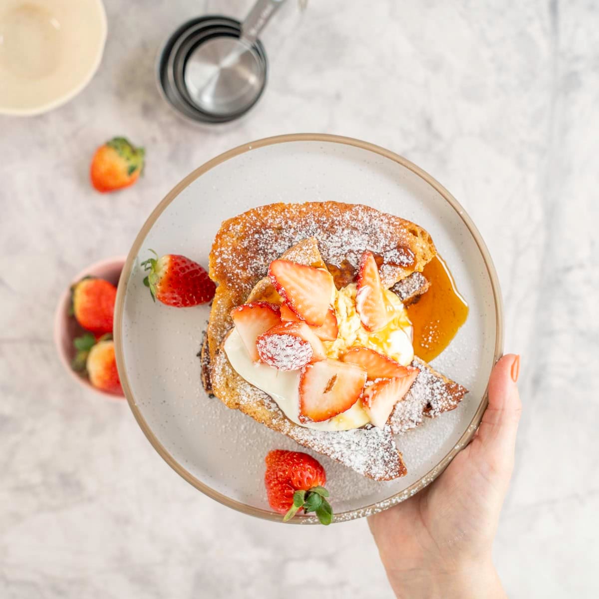 Womans hand holding a plate with stack of french toast with yogurt on top, cut strawberries, icing sugar and maple syrup. Bowl of fresh strawberries and measuring cups below on bench top.