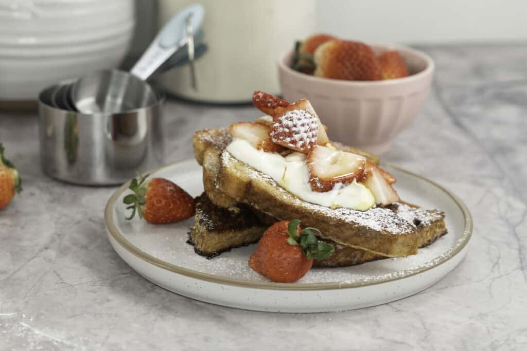 A stack of french toast with yogurt on top, cut strawberries, icing sugar and maple syrup. Bowl of fresh strawberries in small blush pink dish in background and white vases.