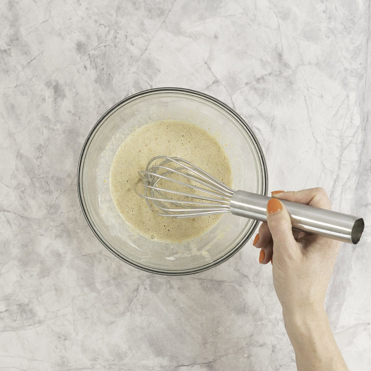 Hand holding a metal whisk, mixing fresh toast mixture inside glass bowl.