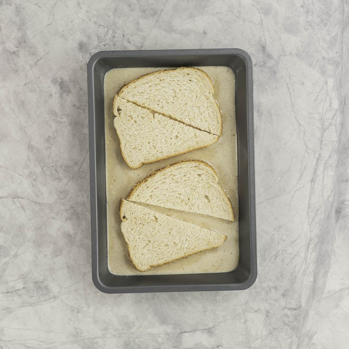 Small rectangle baking tray with french toast mixture inside, two slices of bread cut into triangular pieces.