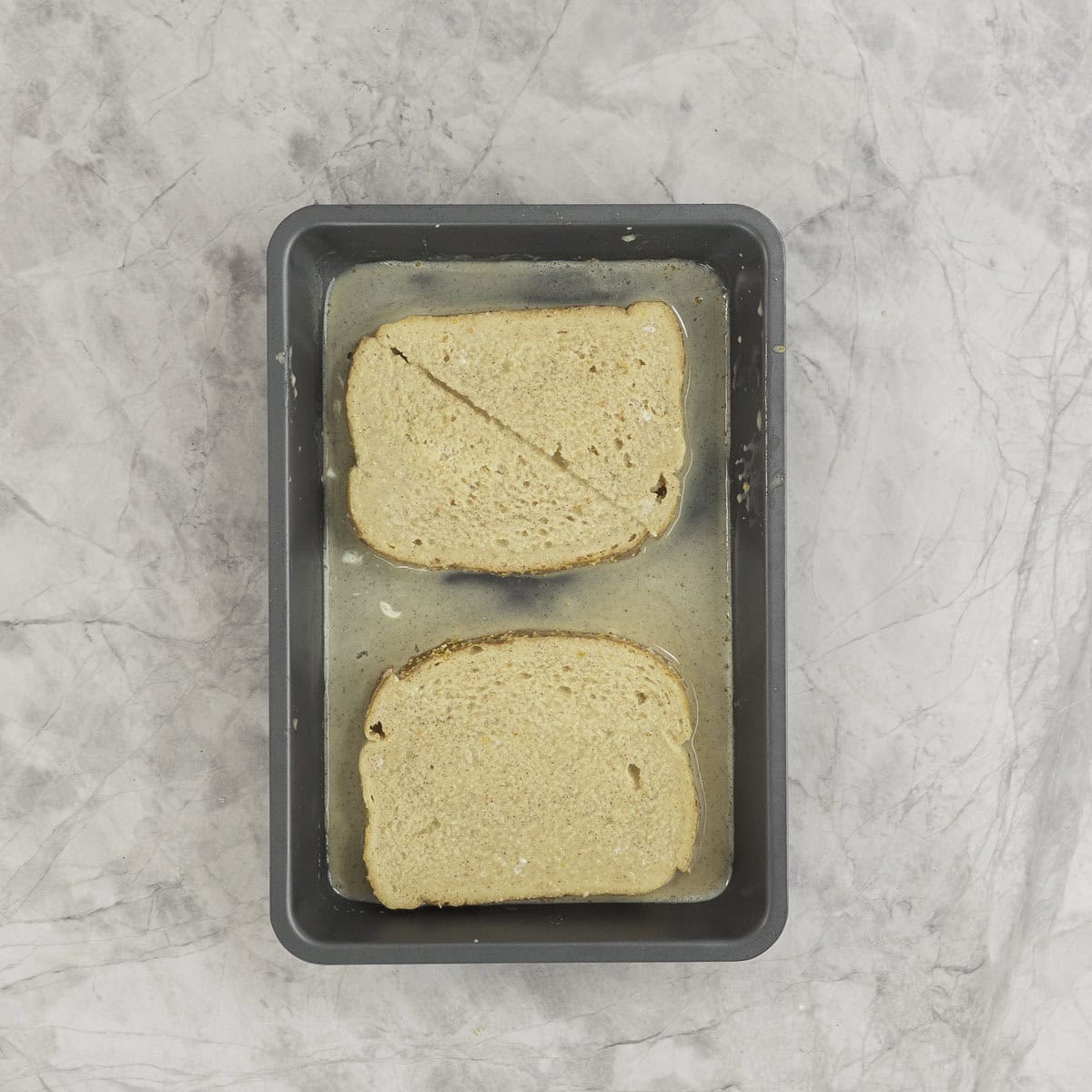 Small rectangle baking tray with fresh toast mixture inside, two slices of bread cut into triangular pieces soaking in the mixture.