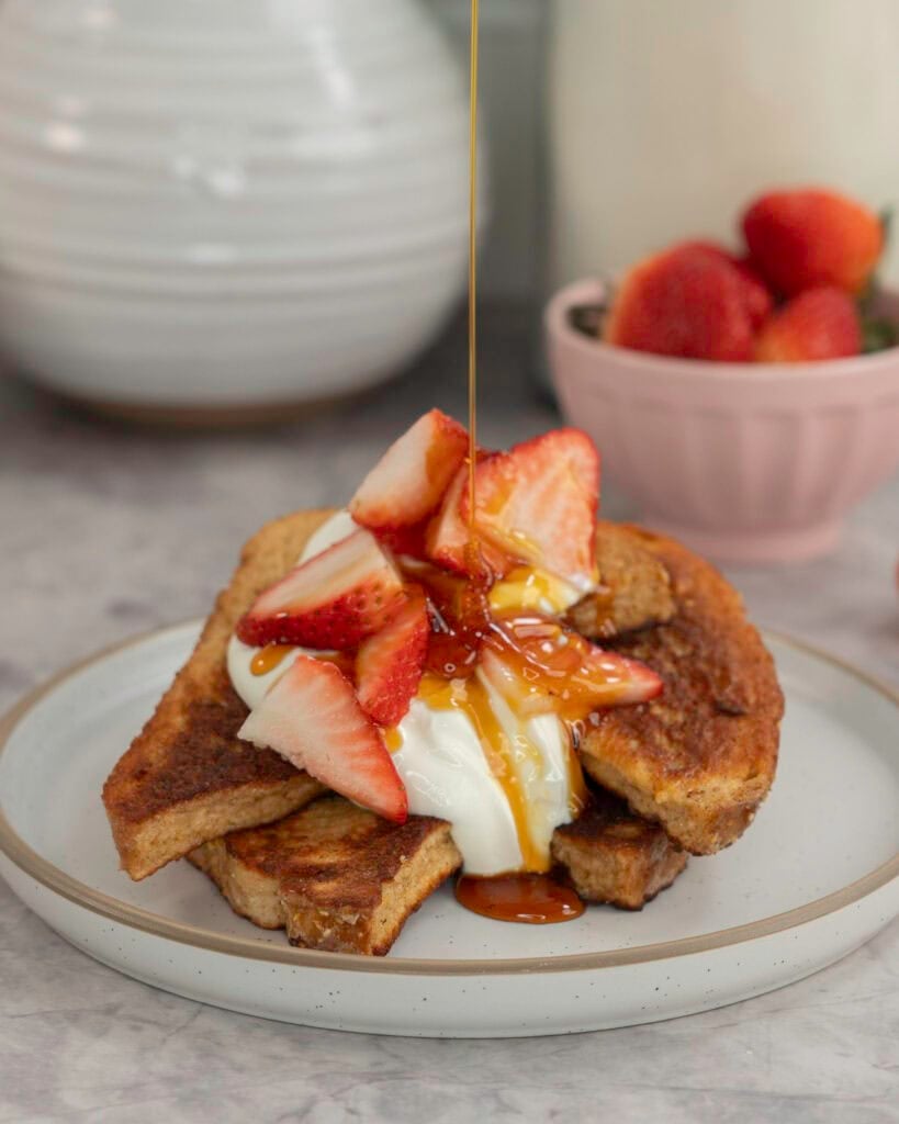 A stack of french toast with yogurt on top and cut strawberries, maple syrup being drizzled on. Bowl of fresh strawberries in small blush pink dish in background and white vases.
