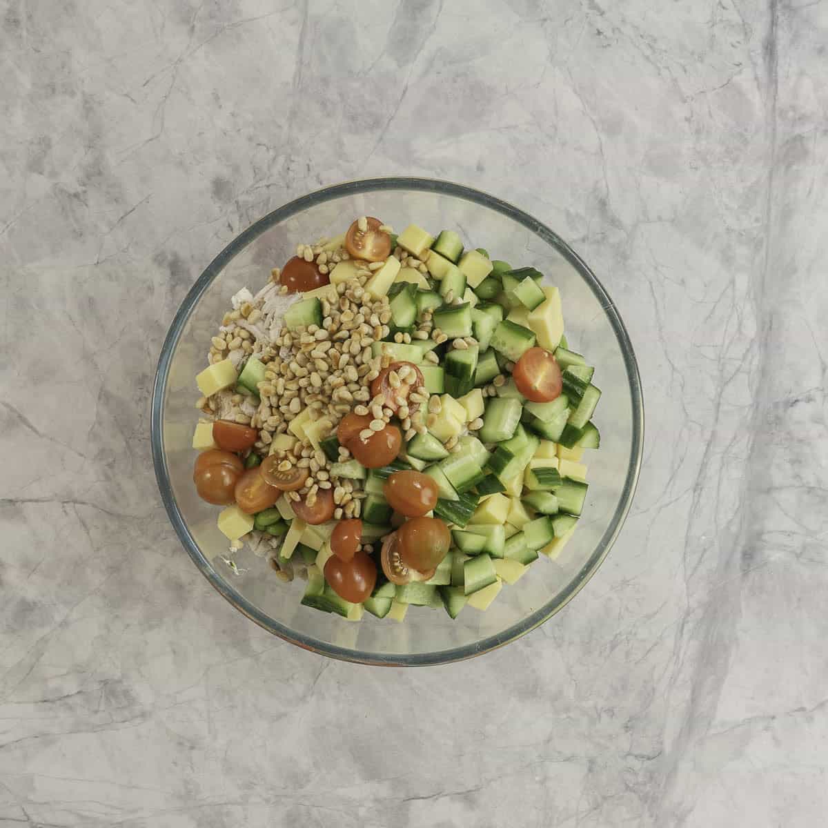 The filings for protein salad in a large glass bowl on benchtop, chicken, pine nuts, cherry tomatoes, cubed cheese and cucumber.