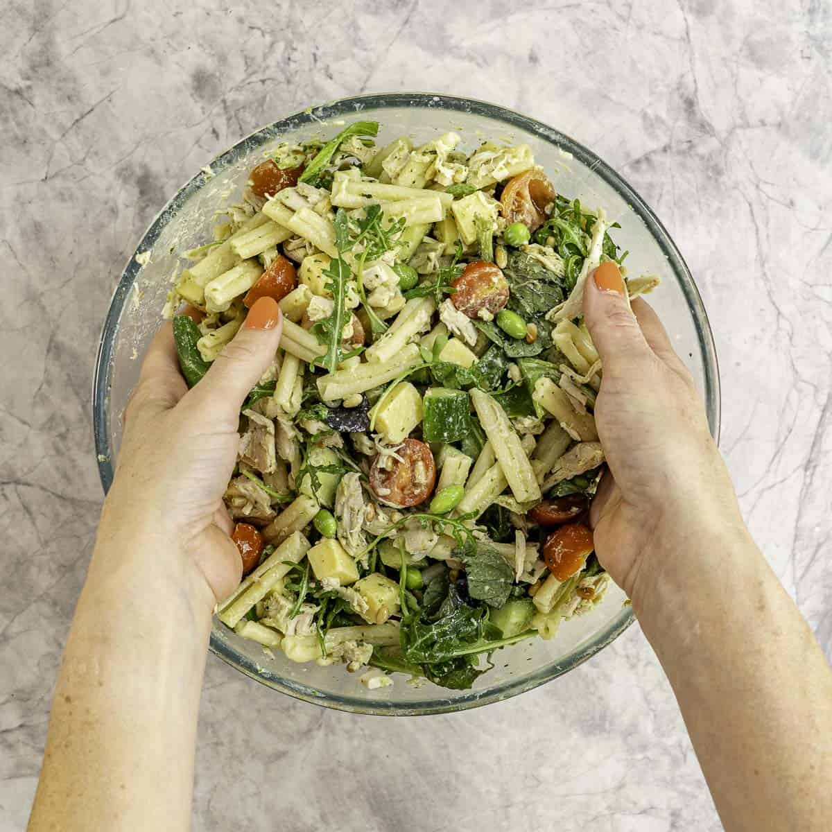 Womans hands inside large glass bowl on marble benchtop tossing together salad inside.