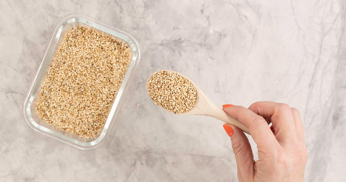 Hand holding a small wooden spoon with puffy quinoa on it, rectangle glass container with quinoa inside on bench top.