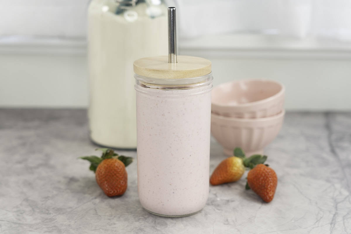 Tall glass with lid and metal straw with pale pink shake inside, fresh strawberries scattered on marble bench top, blush pink bowls in background and large jar of protein.