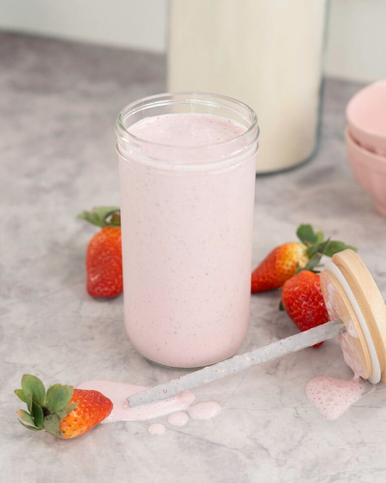 Tall glass with pale pink shake inside, lid and metal straw taken off laying on bench top with shake dripping onto bench, fresh strawberries scattered on marble bench top, blush pink bowls in background and large jar of protein.