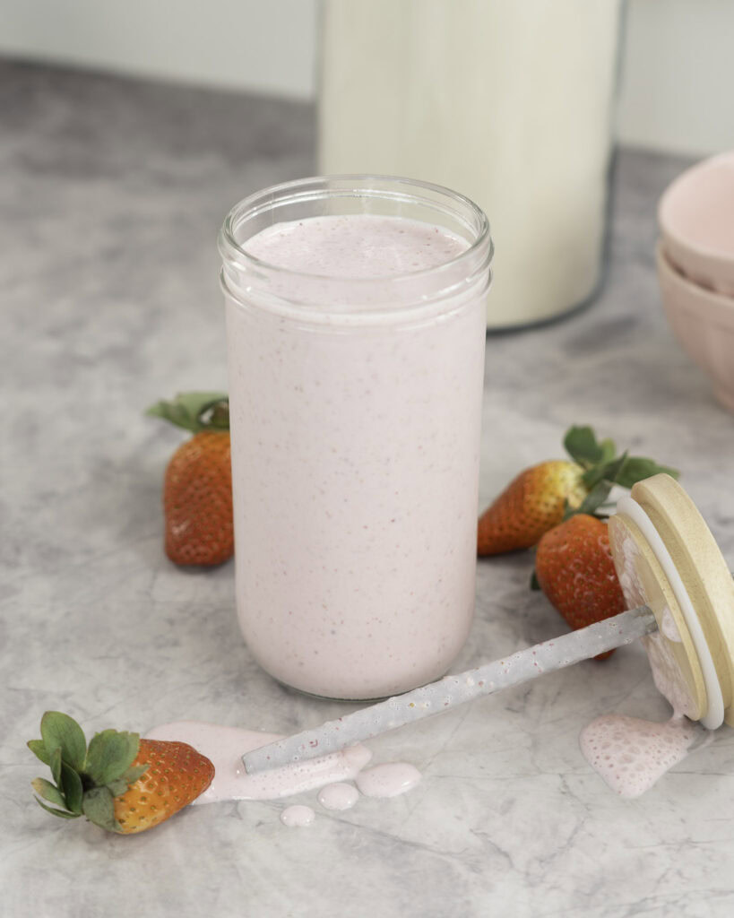 Tall glass with pale pink shake inside, lid and metal straw taken off laying on bench top with shake dripping onto bench, fresh strawberries scattered on marble bench top, blush pink bowls in background and large jar of protein.