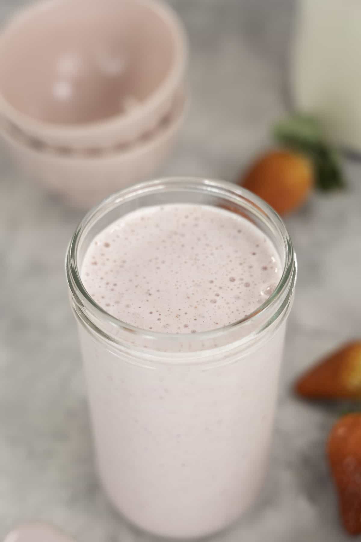 Pale pink smoothie in a tall glass jar, creamy bubbles forming on top, two pink bowls stacked in background and strawberries scattered on bench.