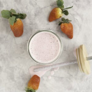 Tall glass with pale pink shake inside, lid and metal straw taken off laying on bench top with shake dripping onto bench, fresh strawberries scattered on marble bench top.