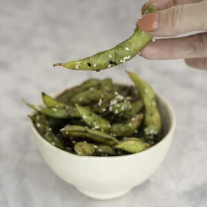 An edamame pod being picked up from a bowl of seasoned edamame.