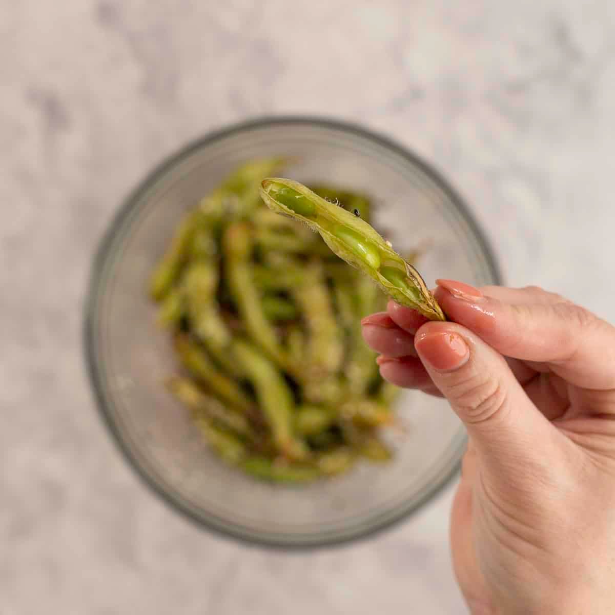 An edamame being held up above a bowl of seasoned edamame with sesame seeds visible.