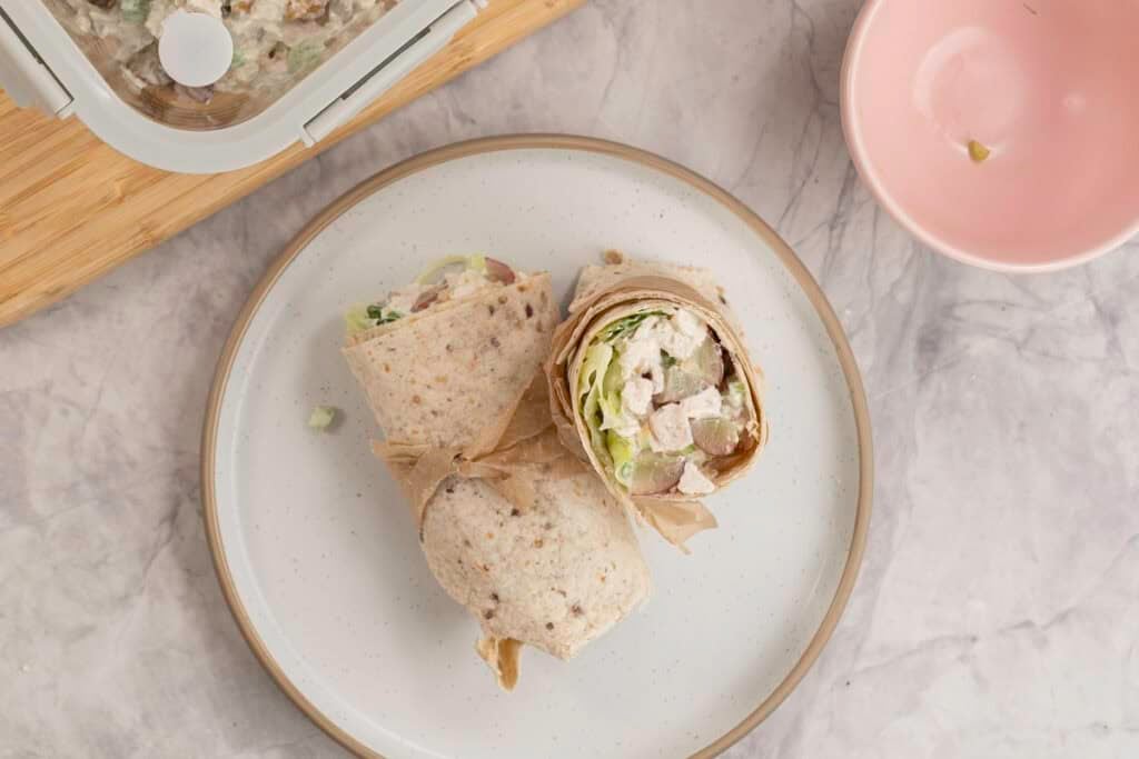 Top down view of a chicken salad wrap on a white plate tied together with string, filling of chicken, grapes and celery with dressing visible. 