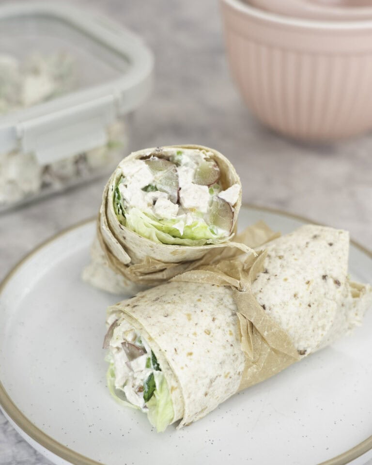 Two halves of a wrap on a plate with a thin piece of brown baking paper tied around each of them. A container with salad in background on benchtop.