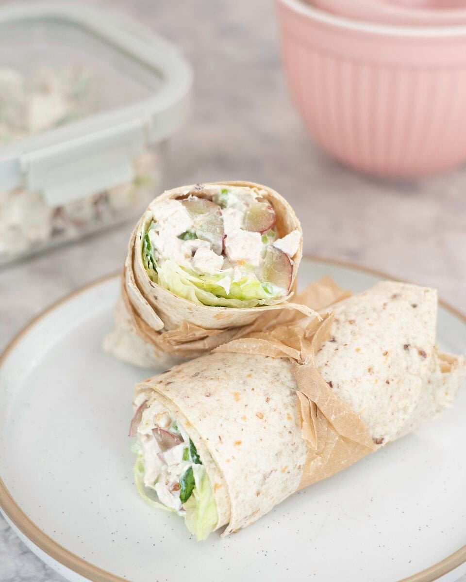 Two halves of a wrap on a plate with a thin piece of brown baking paper tied around each of them. A container with salad in background on benchtop.